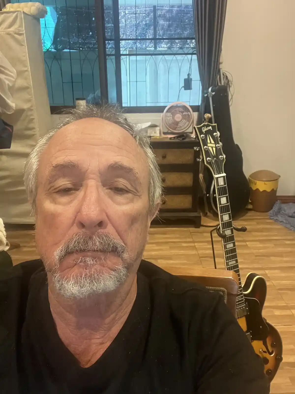 A close-up selfie of an older man with gray hair and a mustache, looking directly at the camera with a tired expression…