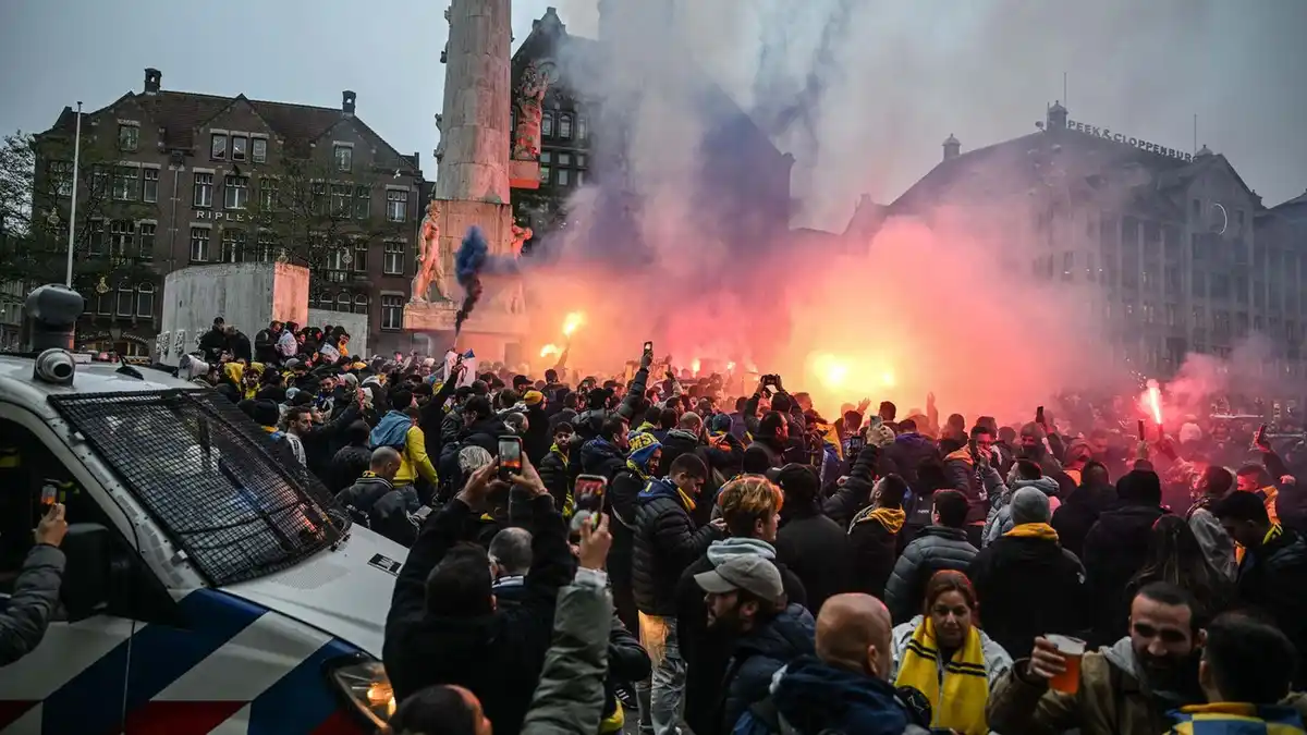A large crowd of people, many wearing yellow and black, are gathered in a city square with red and blue smoke flares…