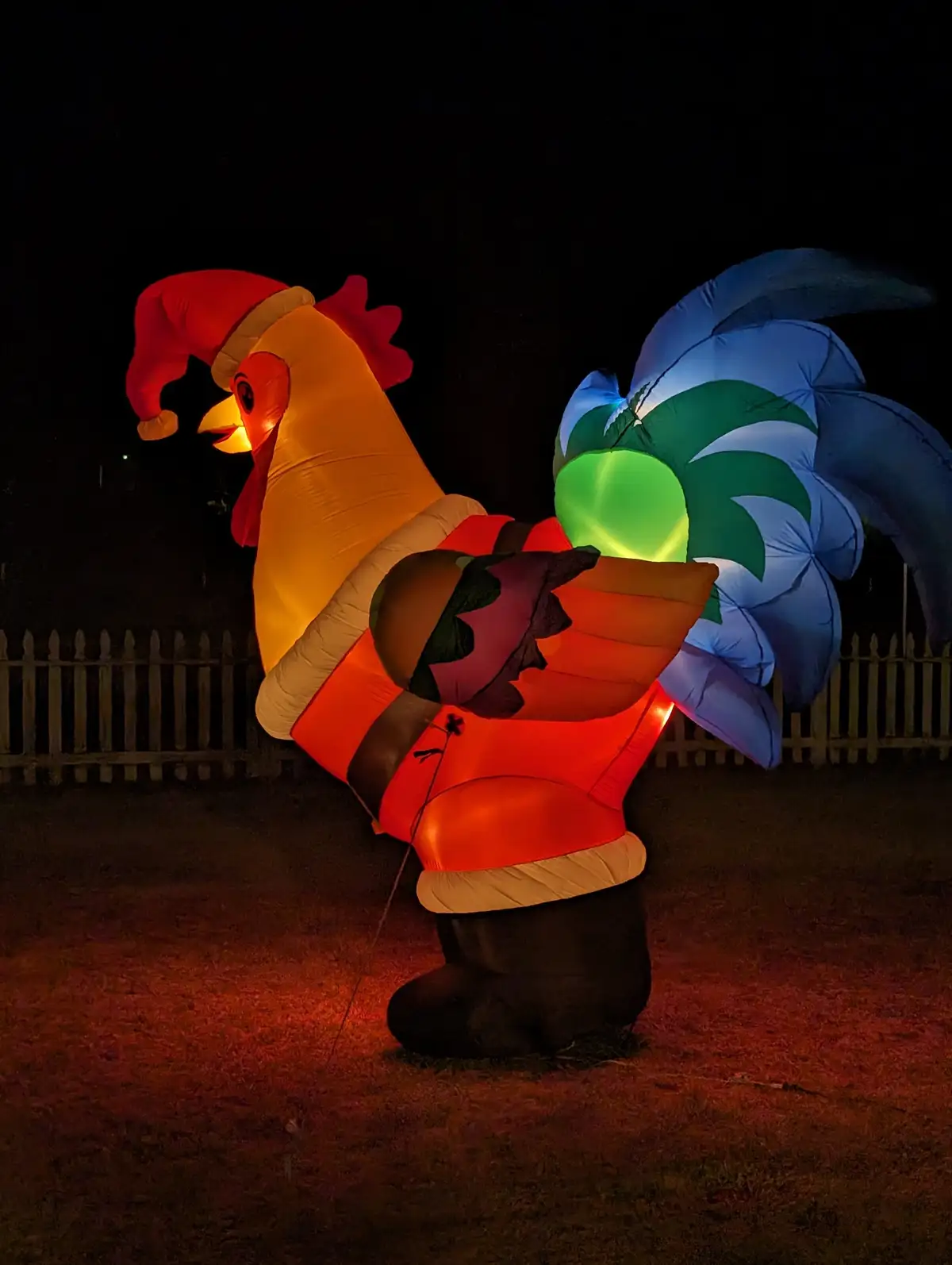A large, illuminated inflatable rooster wearing a red Santa hat and a red Santa suit stands outdoors at night, holding a…