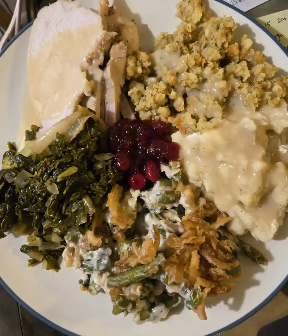 A close-up photo of a white plate piled high with traditional Thanksgiving food: sliced turkey, stuffing, mashed potatoes…