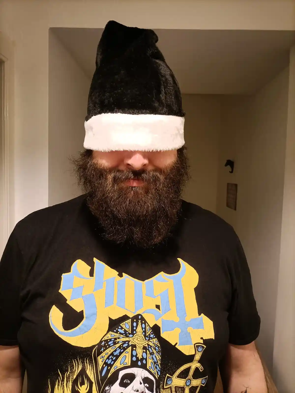 A man with a thick beard and a black t-shirt featuring the 'Ghost' band logo has a black and white Santa hat pulled down…