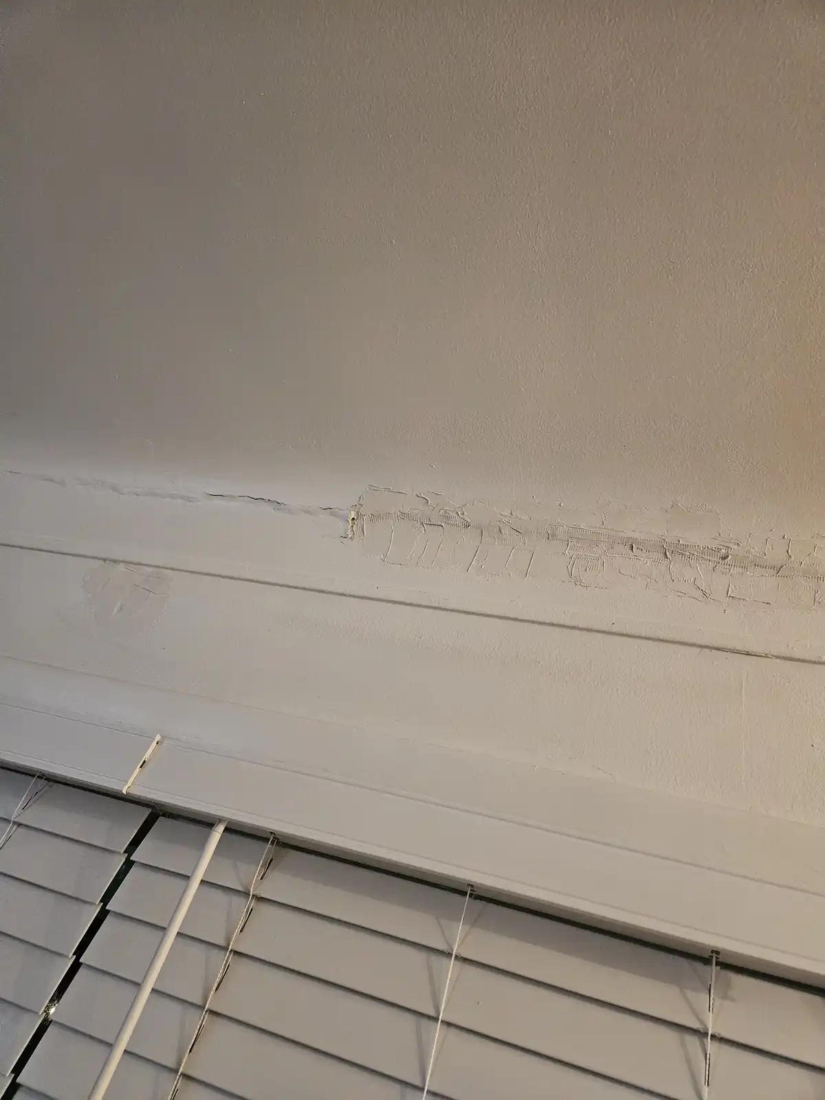 A close-up photo shows a white wall with a visible crack running horizontally near the ceiling, patched with rough, uneven…