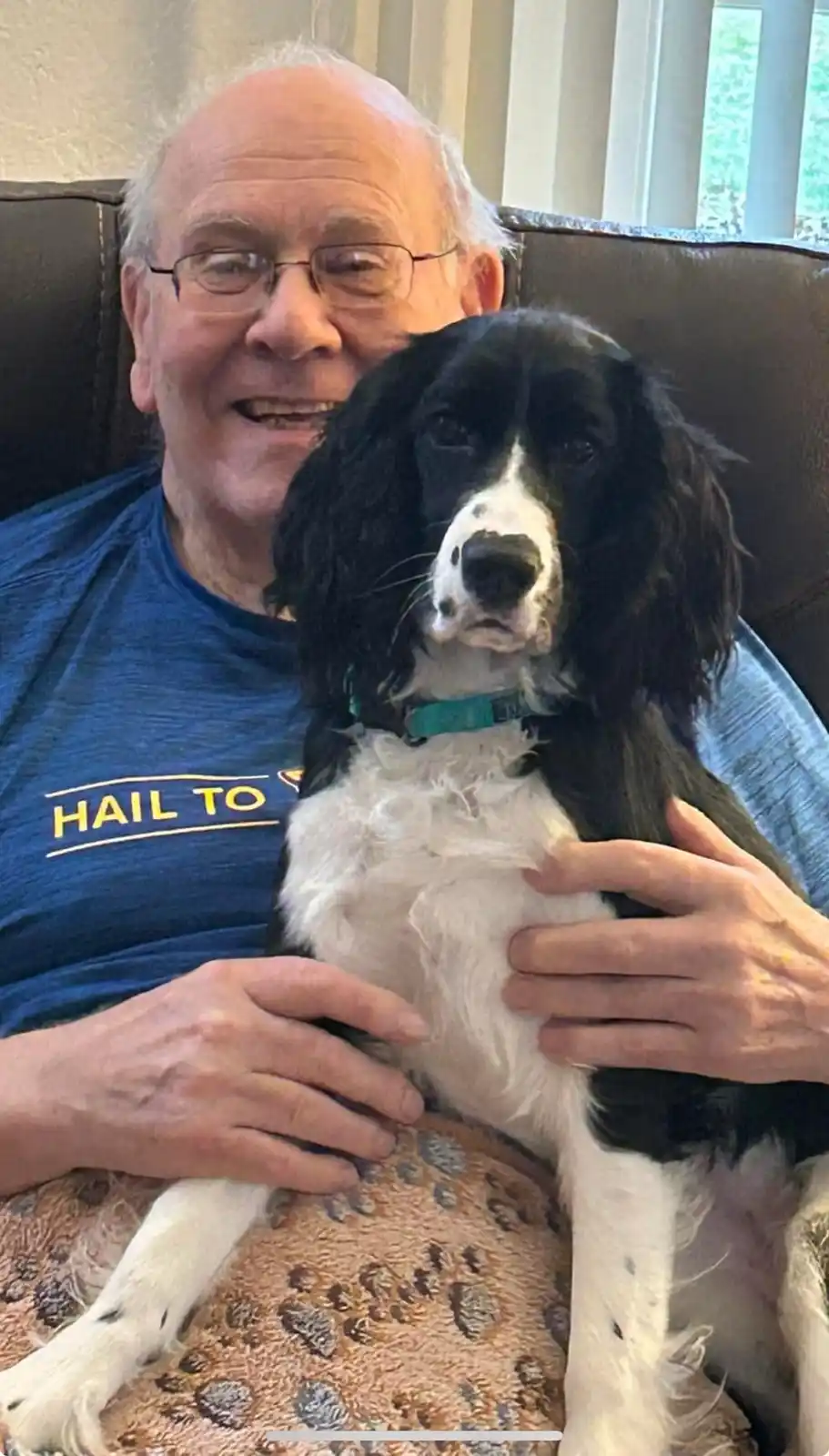 An older man with glasses, wearing a blue t-shirt that says 'HAIL TO', is smiling while holding a black and white dog on his…