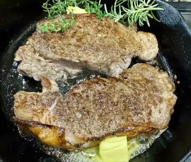 Two seared steaks sizzle in a black cast-iron skillet, topped with fresh rosemary and thyme sprigs and melting butter. The…