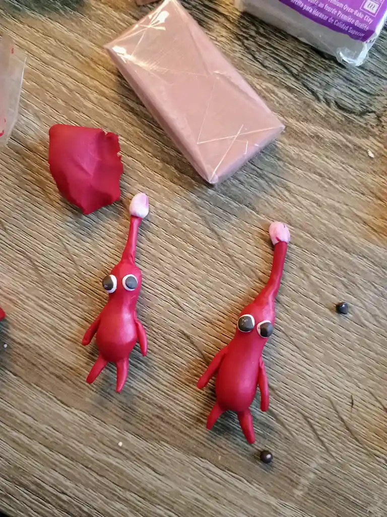 Two small, red clay figures with large googly eyes and long, raised arms sit on a wooden table next to a block of pink clay…