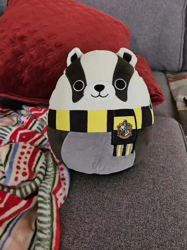 A round, plush badger-shaped Squishmallow toy sits on a gray couch, wearing a yellow and black striped scarf with the…