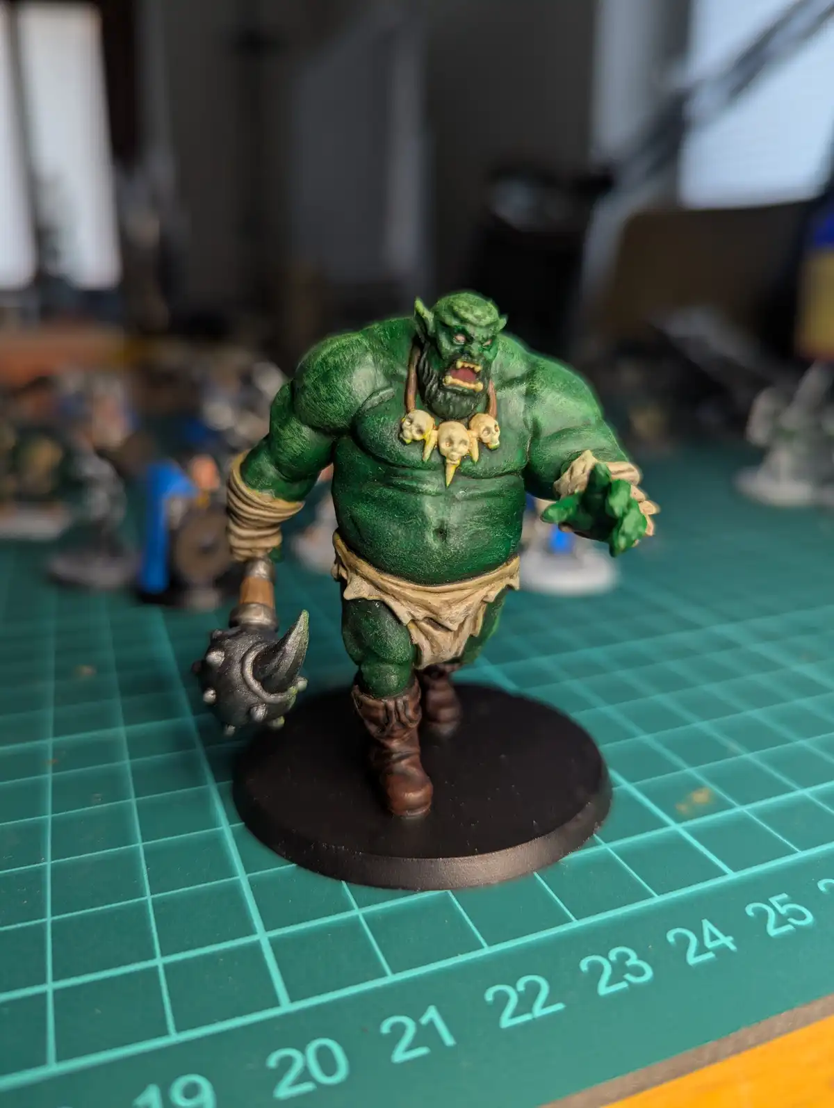 A painted green orc miniature with a spiked mace, skull necklace, and ragged loincloth stands on a black base atop a green…
