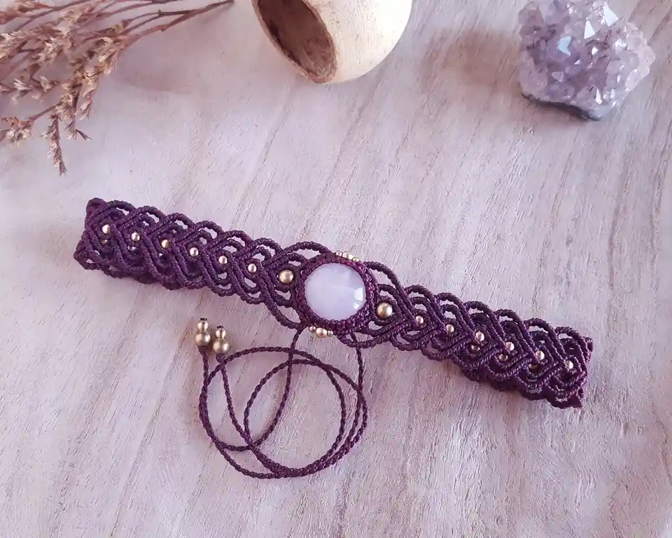 A purple macrame bracelet with a central light purple stone and gold beads lies on a light wooden surface, surrounded by…