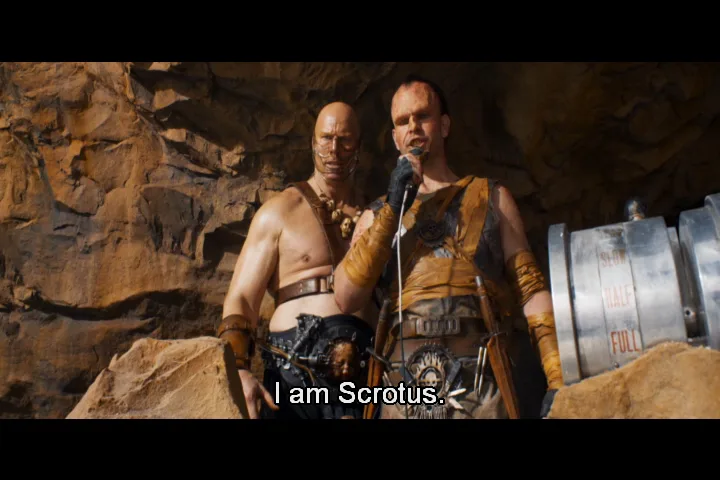 Two muscular men in post-apocalyptic attire stand in a rocky cave, one holding a microphone and speaking. The man speaking…