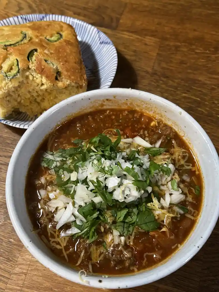 A close-up photo shows a white bowl filled with chili topped with shredded cheese, chopped white onions, and fresh green…