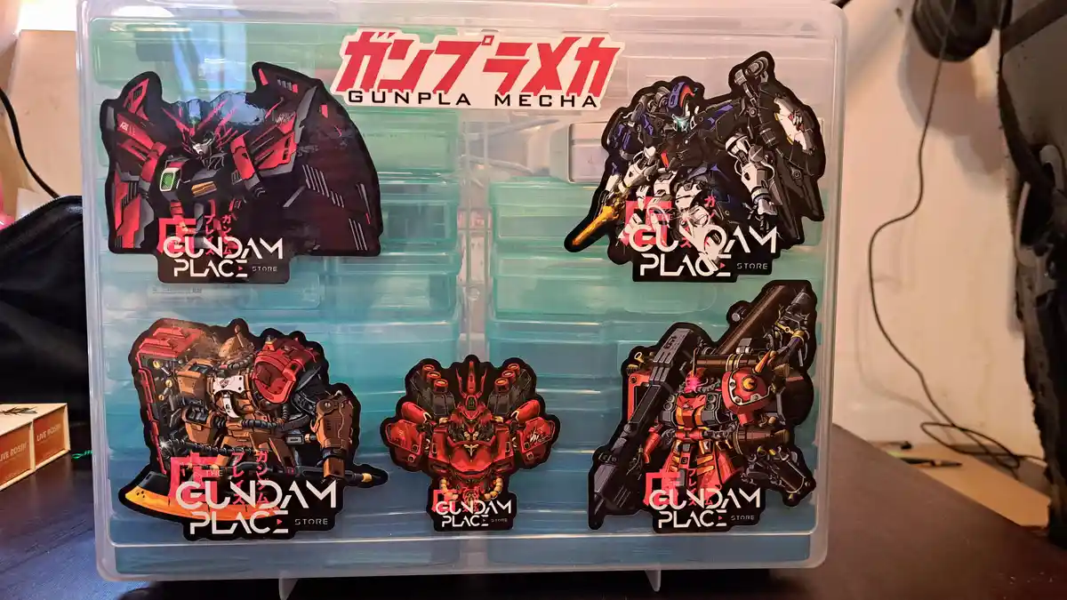 A clear plastic storage bin labeled 'ガンプラメカ GUNPLA MECHA' is decorated with five large, colorful Gundam-themed stickers…