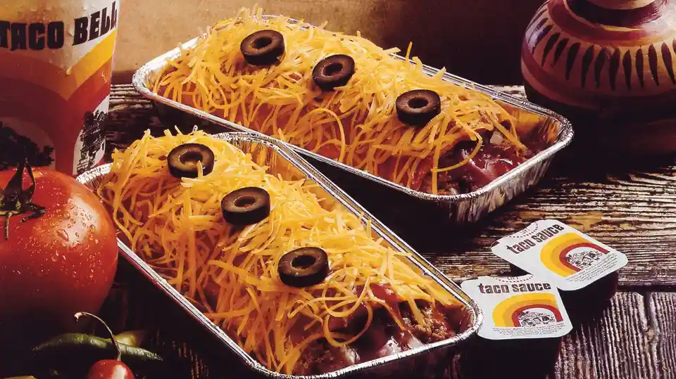 Two aluminum trays of Taco Bell-style tacos topped with shredded cheese and black olives arranged to look like faces, with a…
