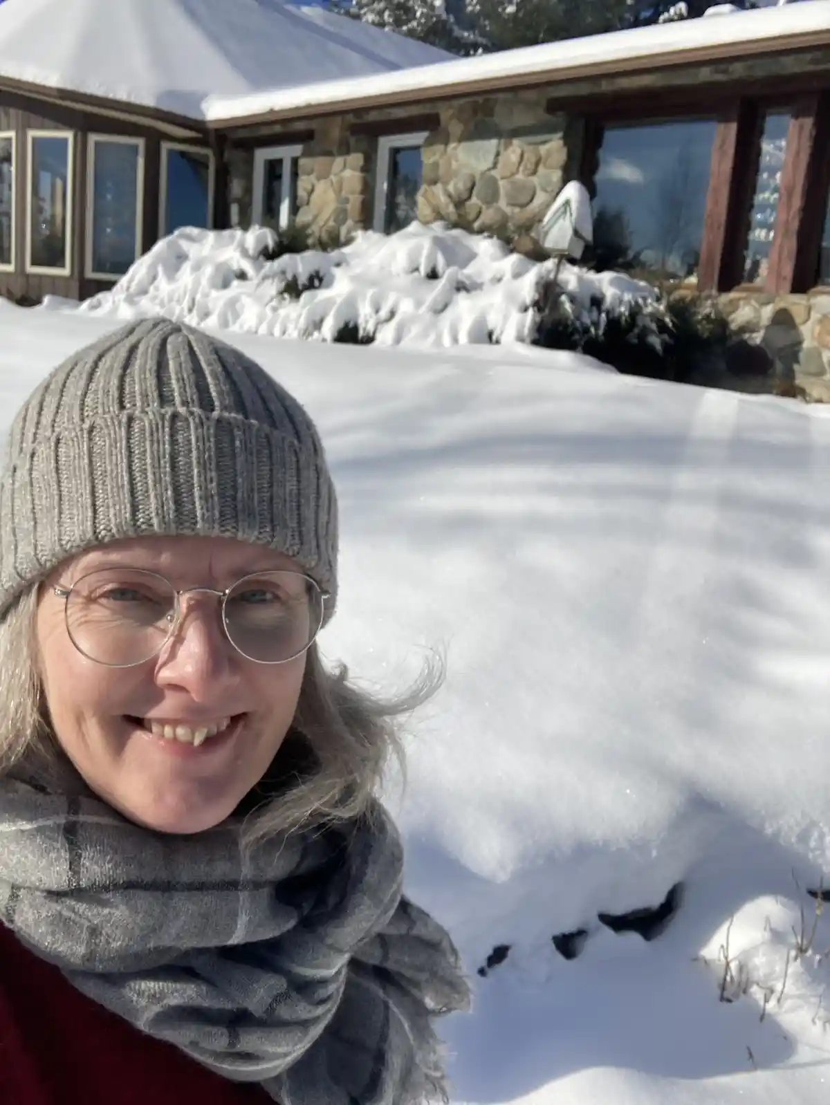 A woman with gray hair, wearing a gray knit beanie, round glasses, and a gray scarf, smiles at the camera while standing in…