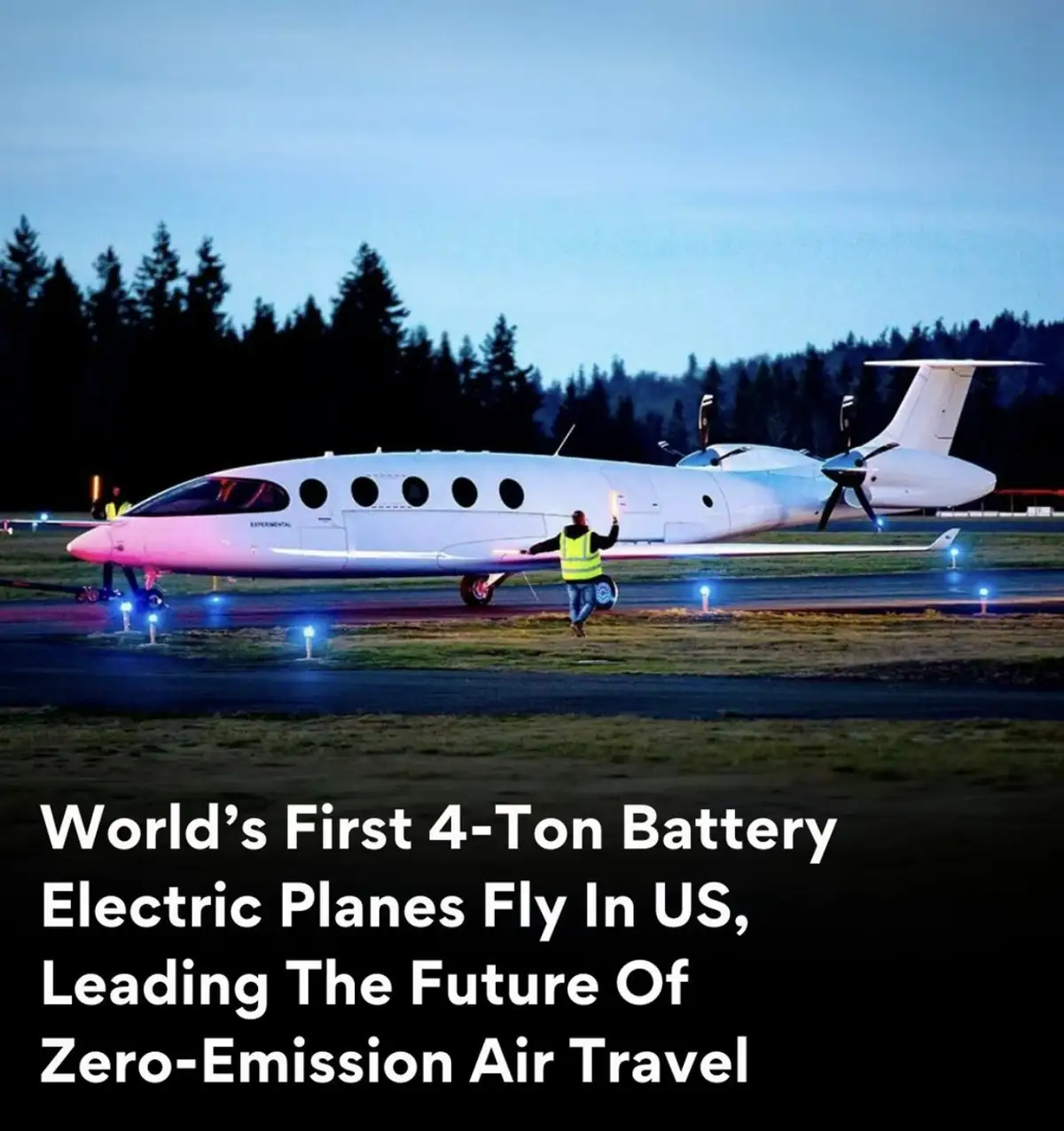 A white experimental electric airplane is parked on an airport tarmac at dusk, with a ground crew member in a yellow vest…