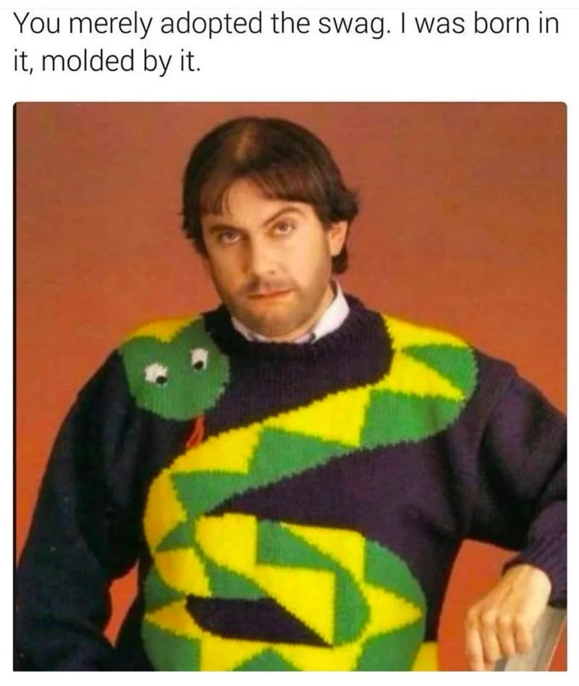 A man with dark hair and a mustache stares intensely at the camera while wearing a dark purple sweater featuring a large…