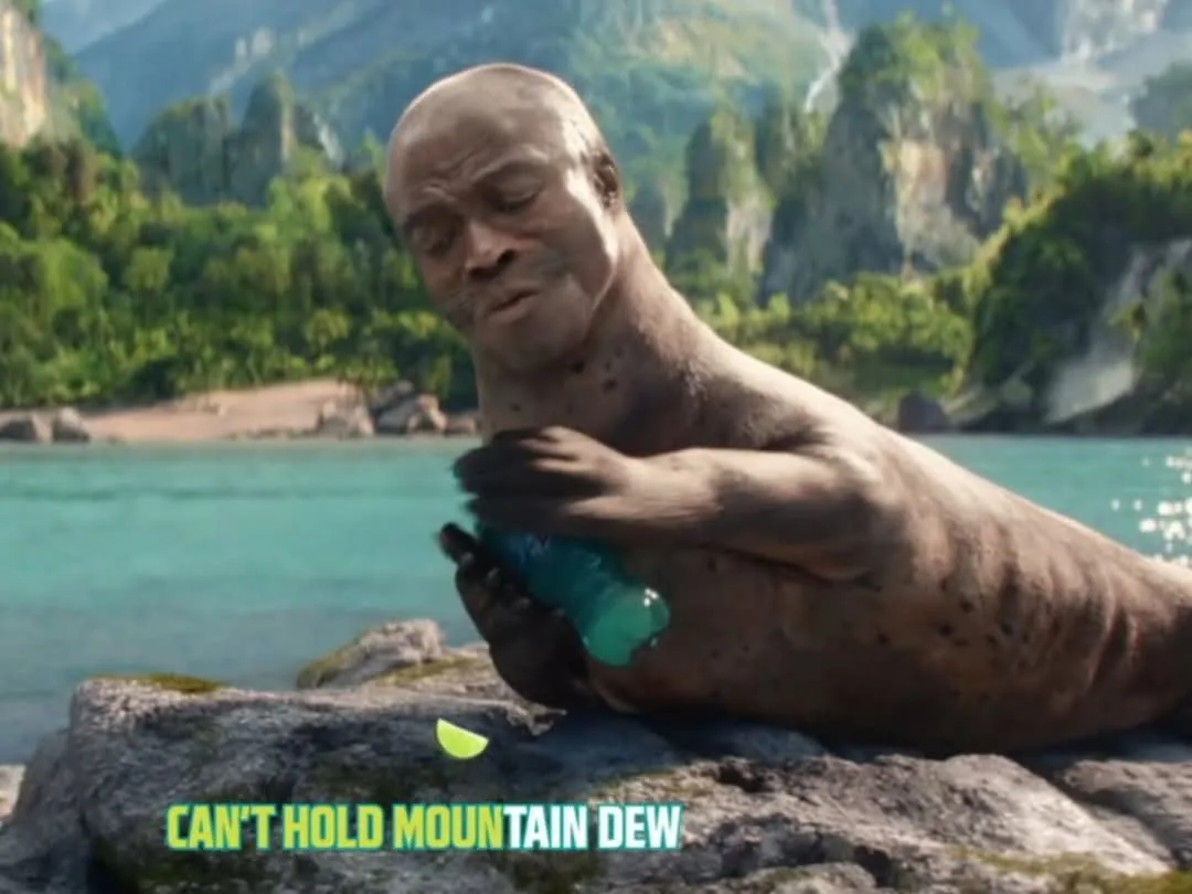 A bald, muscular man with a seal-like body is sitting on a rock by a turquoise lake, struggling to hold a green Mountain Dew…