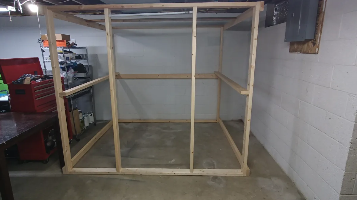A wooden frame structure resembling a closet stands in a basement or garage, with white cinder block walls and concrete…