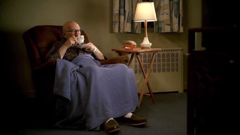 An elderly man with glasses sits in a recliner, wrapped in a blue blanket, sipping tea from a cup. A lamp and small table…