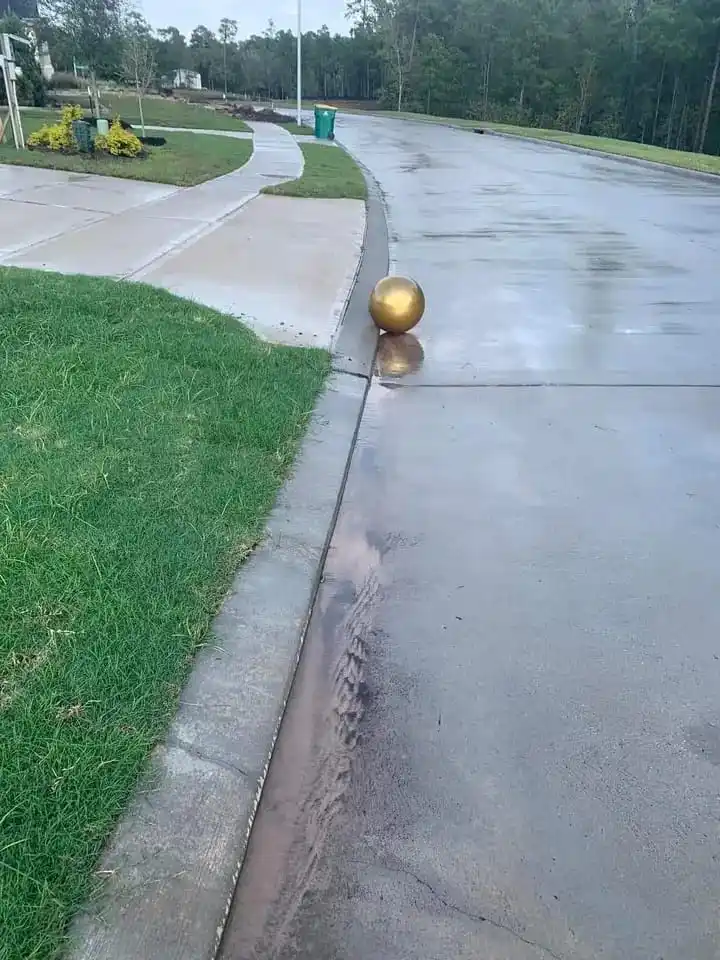 A shiny gold ball rests precariously balanced on the edge of a wet concrete driveway curb, with grass and a sidewalk visible…