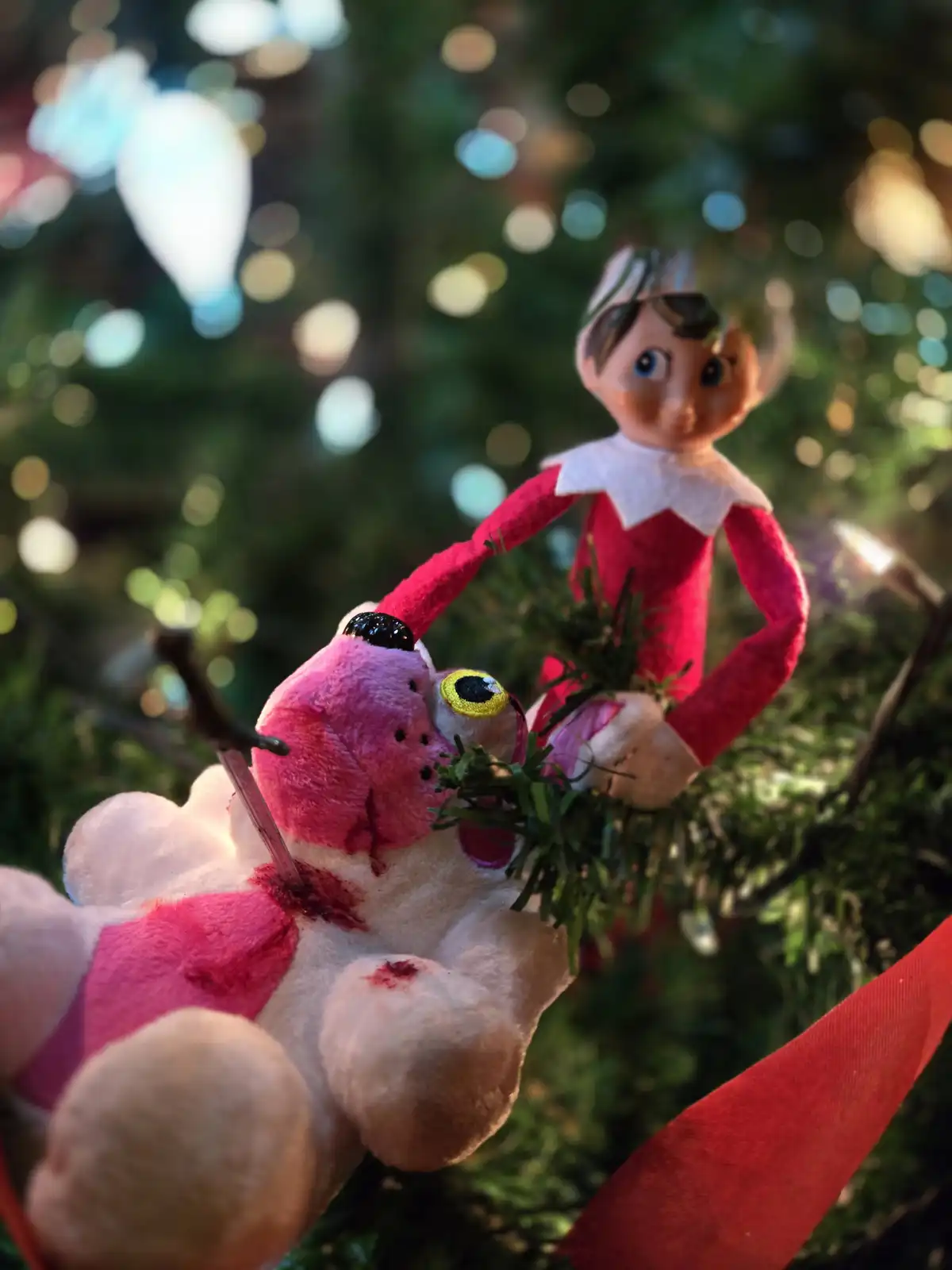 A plush pink and white teddy bear with fake blood and a single yellow eye is pinned to a Christmas tree branch by an elf in…