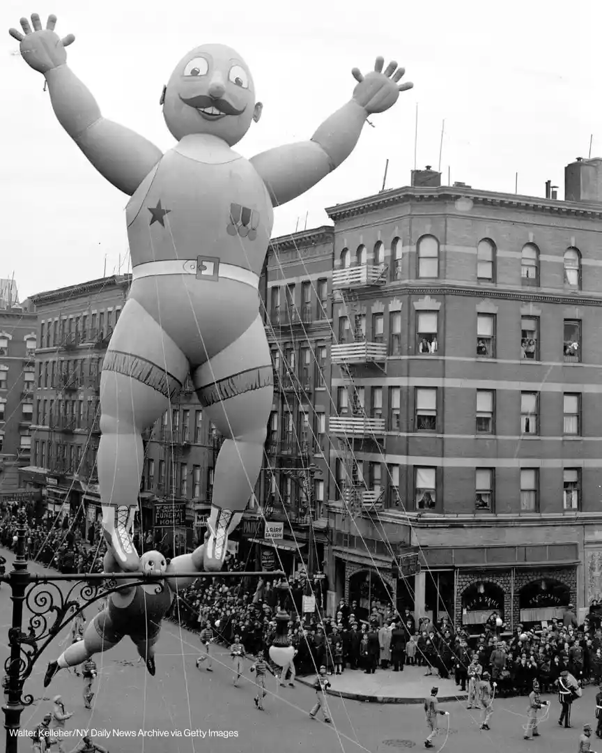 A black-and-white photo shows a giant, cartoonish inflatable man with a mustache, star, and medals, floating above a crowded…