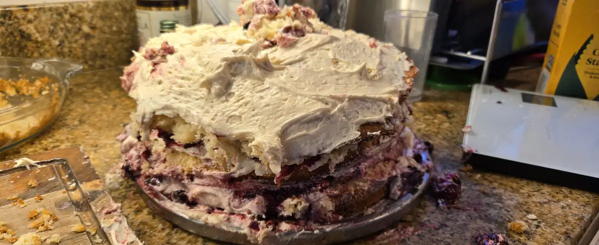 A messy, multi-layered cake with white frosting and purple berry filling sits on a metal cake pan on a granite countertop…