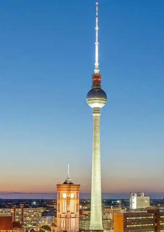 A tall, illuminated tower with a spherical observation deck stands against a twilight sky over a cityscape. The overlaid…