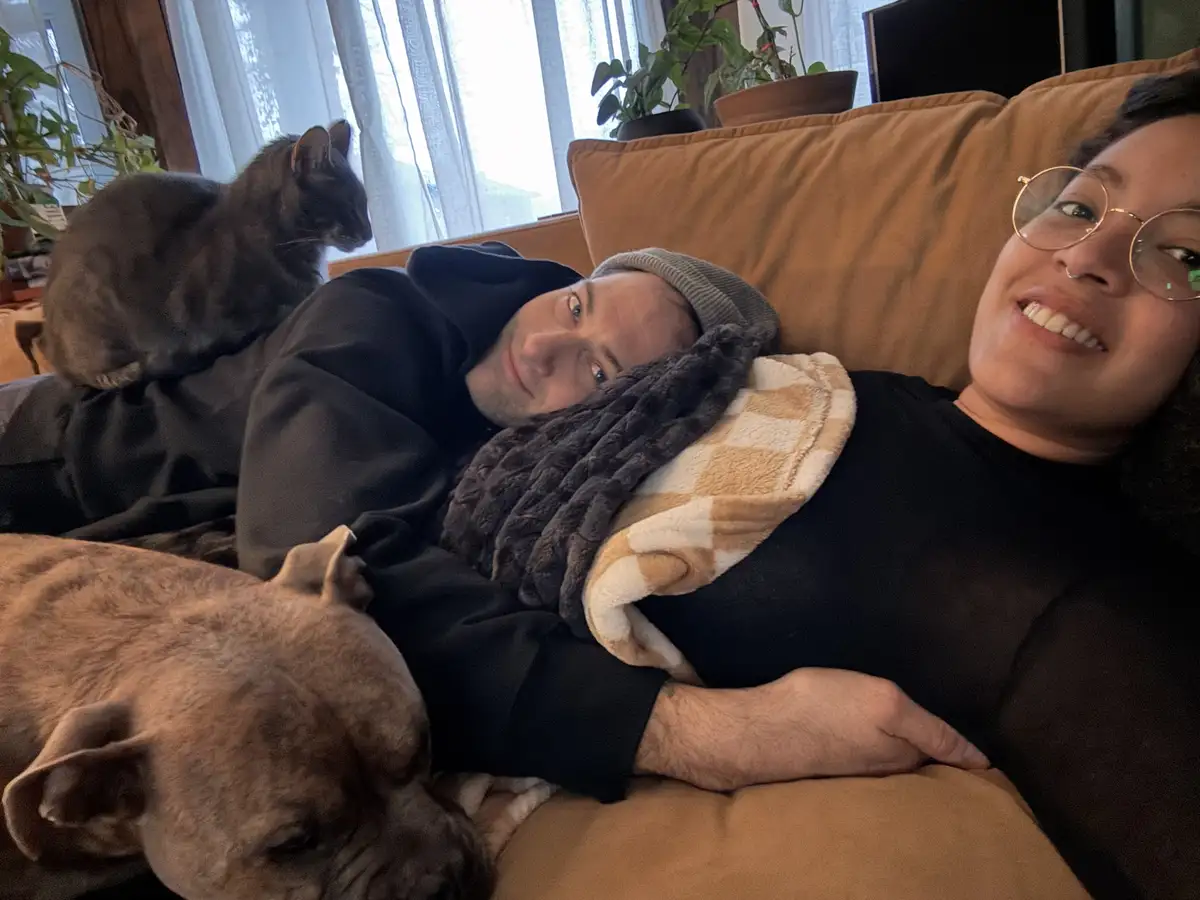 A man and woman are lying on a brown couch, smiling at the camera, with a black cat sitting on the man's back and a brown…