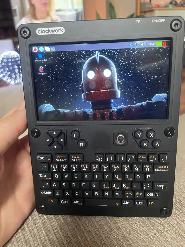 A hand holds a black 'clockwork' branded handheld device with a full keyboard and screen displaying a cartoon robot with…
