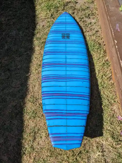 A blue surfboard with purple stripes lies on dry grass next to a wooden deck. The overlaid text reads 'i'm not a surfer, i'm…
