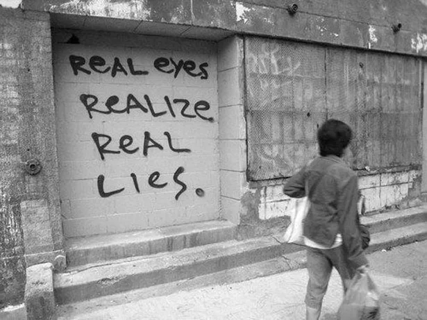 A black-and-white photo shows a person walking away from a boarded-up doorway with graffiti that reads 'REAL EYES REALIZE…