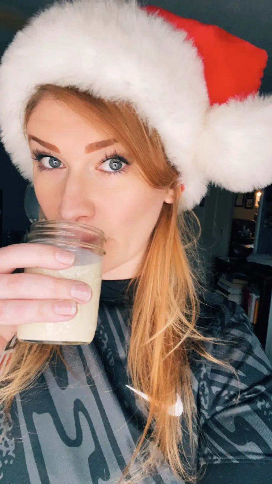 A woman with red hair and blue eyes wears a red and white Santa hat while holding a mason jar filled with a light-colored…