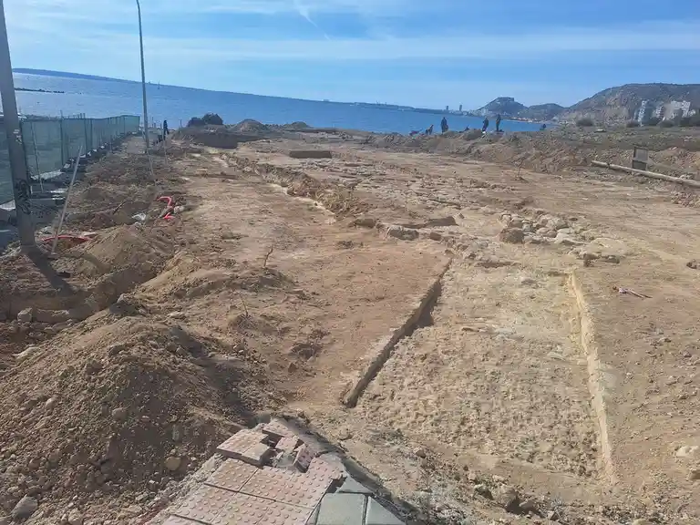 An outdoor construction site near a body of water shows exposed ancient stone foundations and walls, with piles of dirt and…