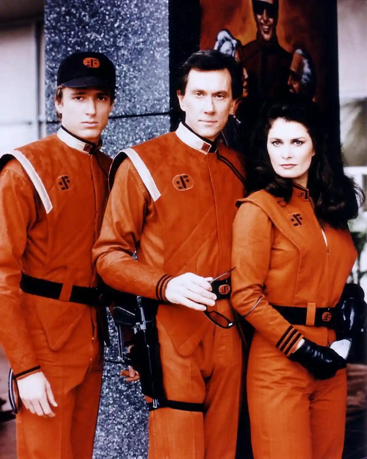 Three actors in orange Star Trek uniforms stand together, looking serious. The man in the center holds glasses, the woman on…