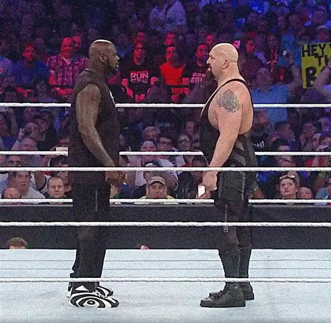 Two muscular wrestlers face off in a ring with a crowd behind them. The wrestler on the left wears black pants and…