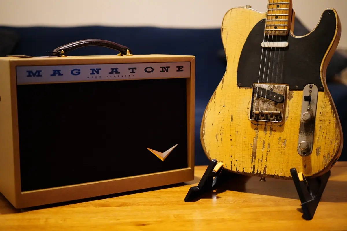 A vintage-style yellow electric guitar with a heavily worn, cracked finish sits on a stand next to a tan Magnatone 'High…