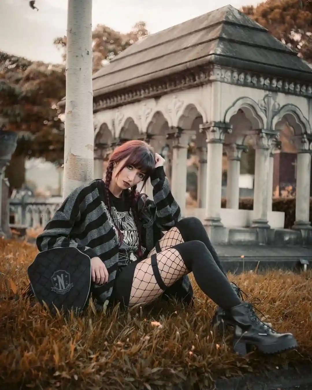 A young woman with red hair in braids sits on dry grass in a cemetery, wearing a black-and-gray striped sweater, fishnet…