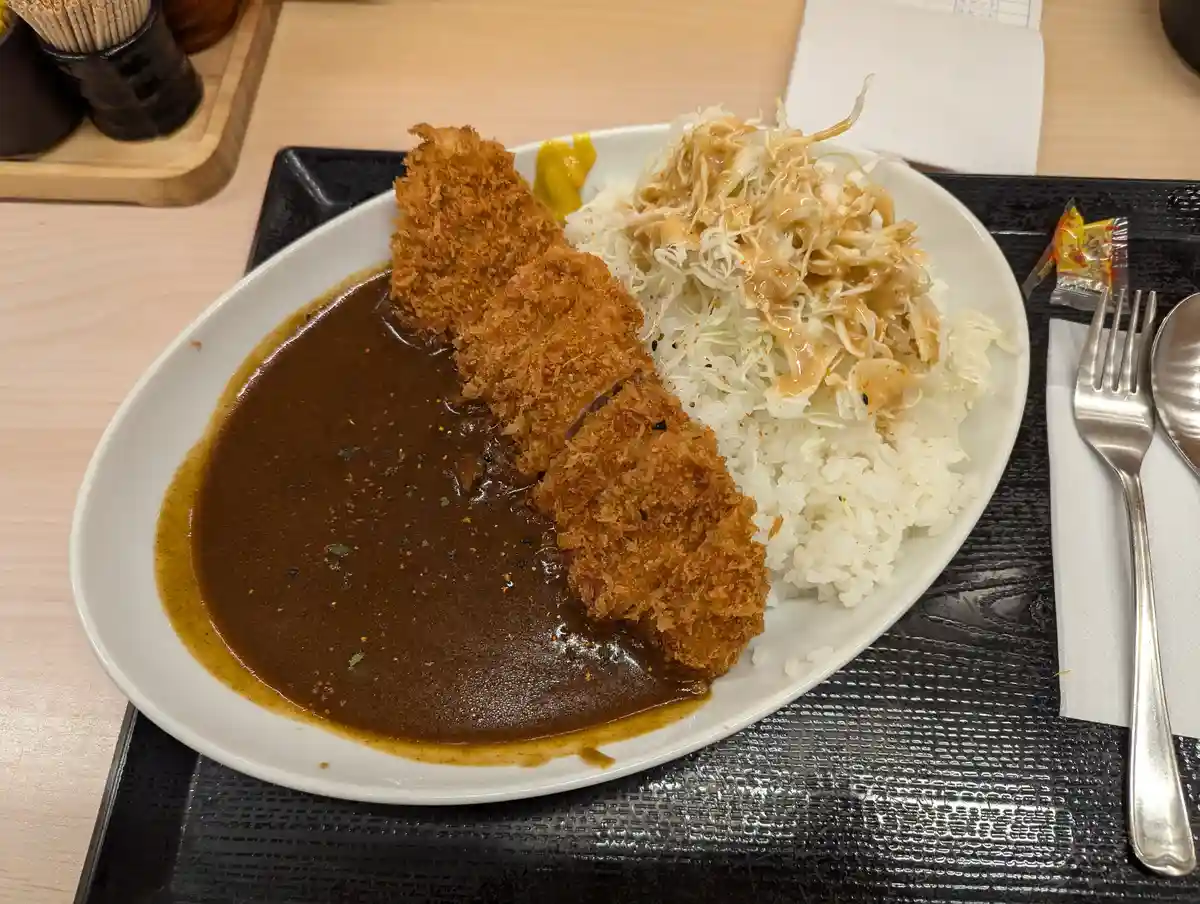 A close-up photo of a Japanese katsu curry meal on a white plate, featuring a breaded and fried pork cutlet (katsu)…