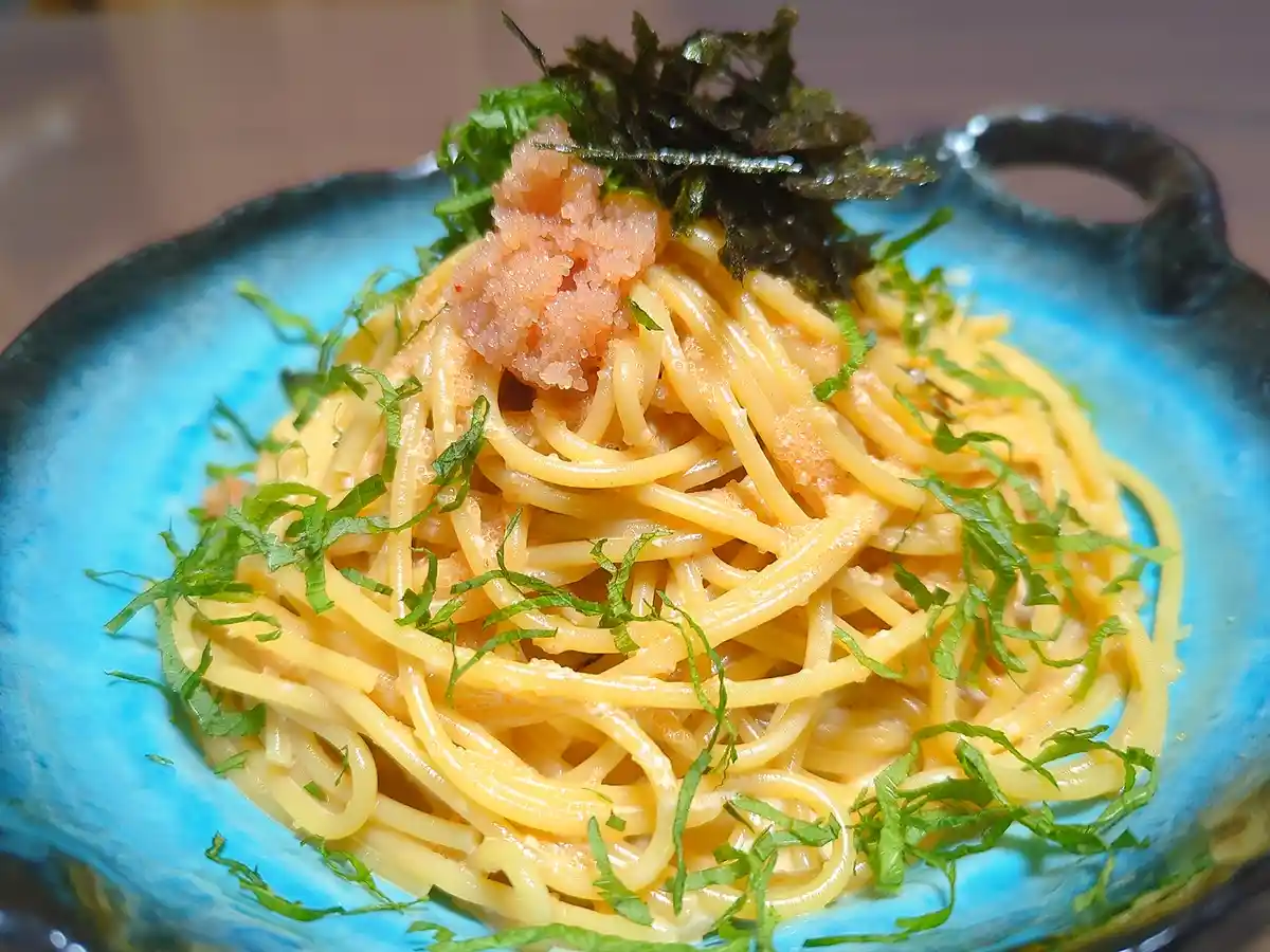 a close-up photo of spaghetti in a blue bowl, topped with pink tobiko, green shredded herbs, and dark nori seaweed flakes…