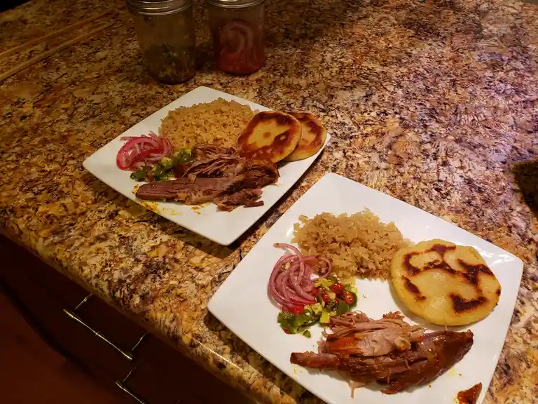 Two white plates on a granite countertop each hold a meal of shredded meat, rice, pickled onions, and a small salsa. The…