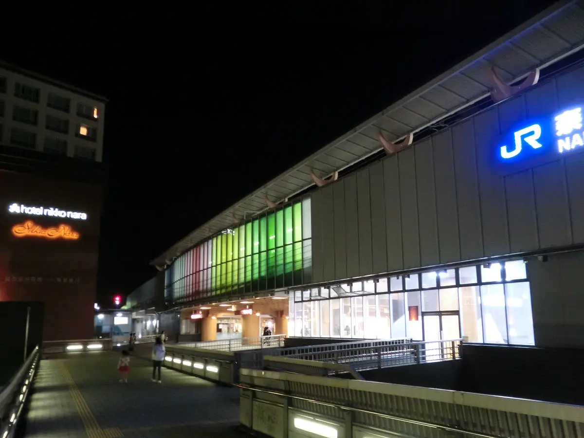 A nighttime photo of JR Nara station, featuring its illuminated facade with a rainbow-colored light strip and the glowing…
