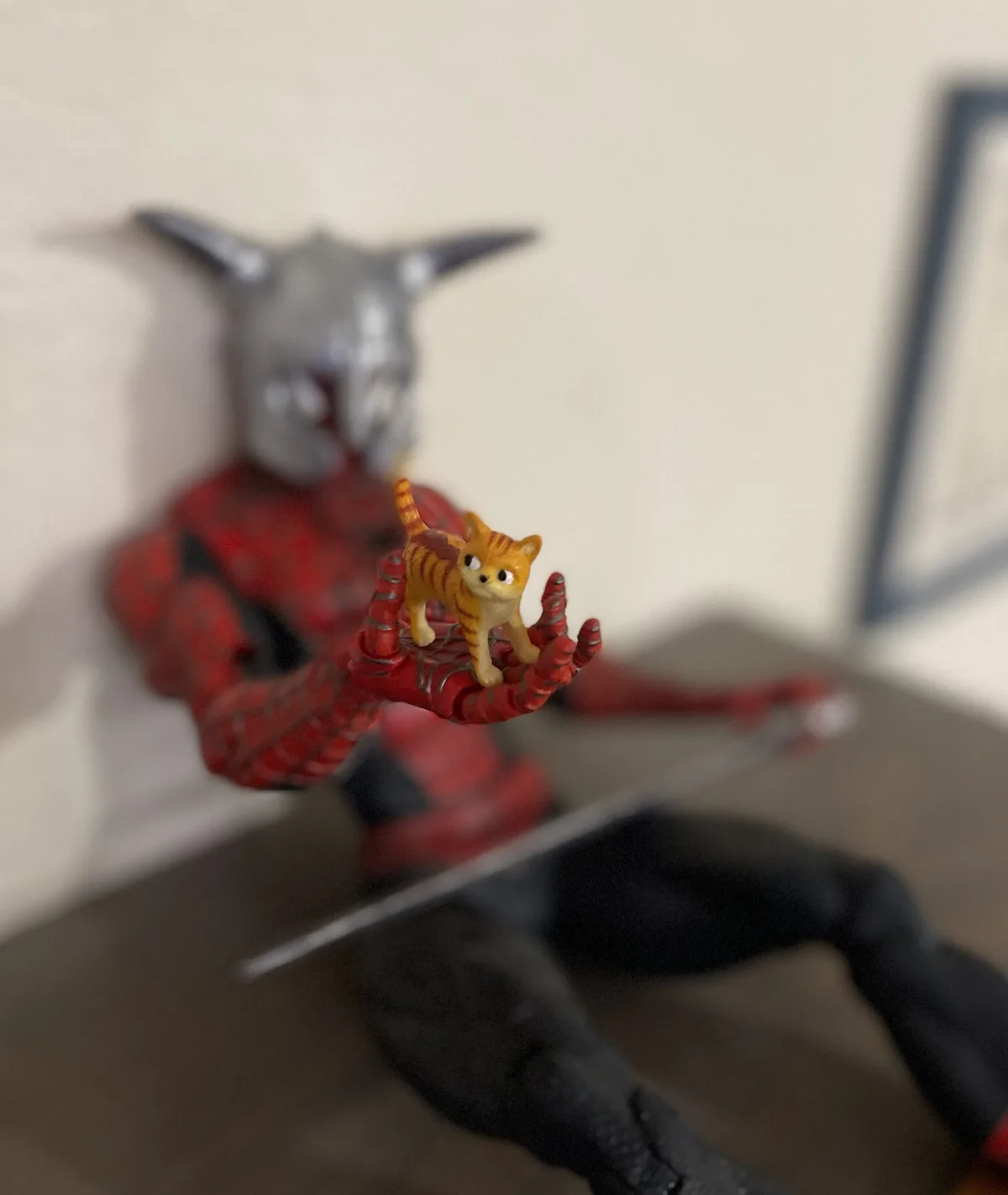 A close-up photo shows a small orange tabby cat figurine standing on the open palm of a Spider-Man action figure. The…