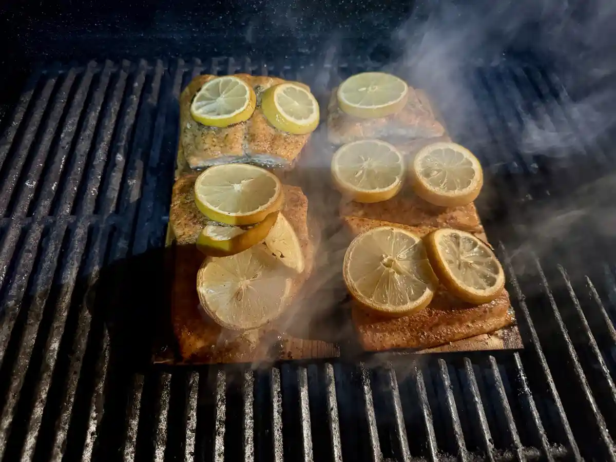 Salmon fillets are cooking on a grill, each topped with multiple lemon slices, with steam rising from the hot grates. The…