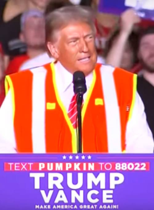 A man wearing a bright orange and white striped safety vest stands at a podium with a microphone. Behind him, a crowd is…