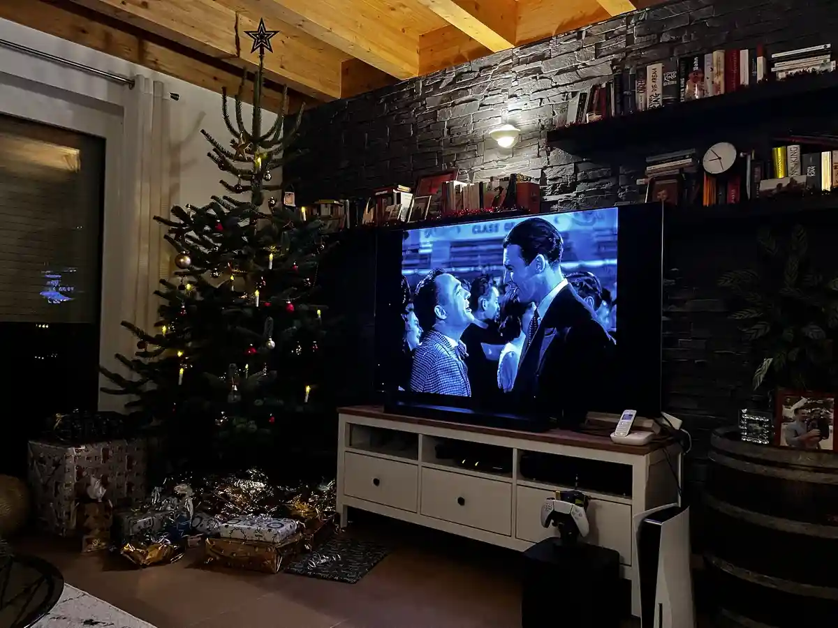 A cozy, dimly lit living room features a decorated Christmas tree with presents underneath, a stone wall with bookshelves…