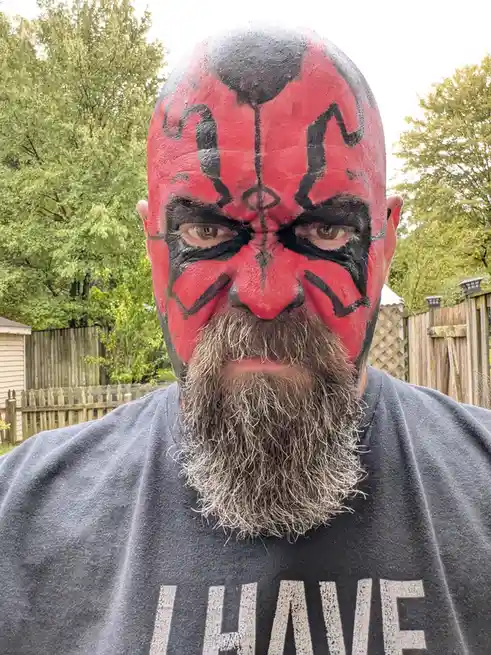 A man with a full beard and bald head is shown in a selfie, his face painted red and black to resemble the character Darth…