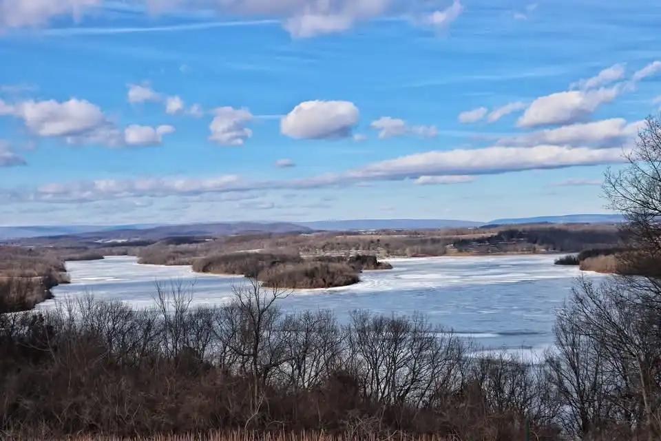 A wide landscape photo shows a frozen lake under a bright blue sky with scattered white clouds, surrounded by bare winter…