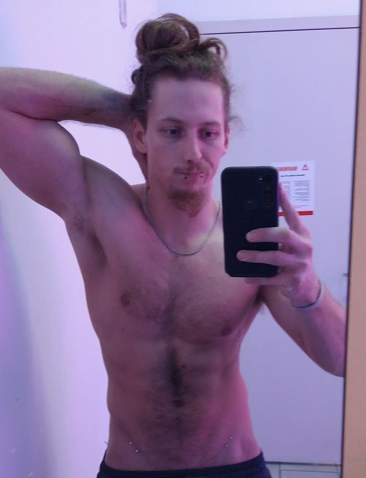 A shirtless man with a bun hairstyle and visible tattoos takes a mirror selfie with a black phone, flexing his arm while…