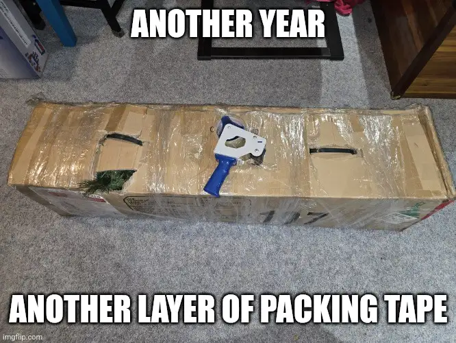 A long cardboard box lies on a carpeted floor, heavily wrapped in clear packing tape with visible tears. A blue and white…