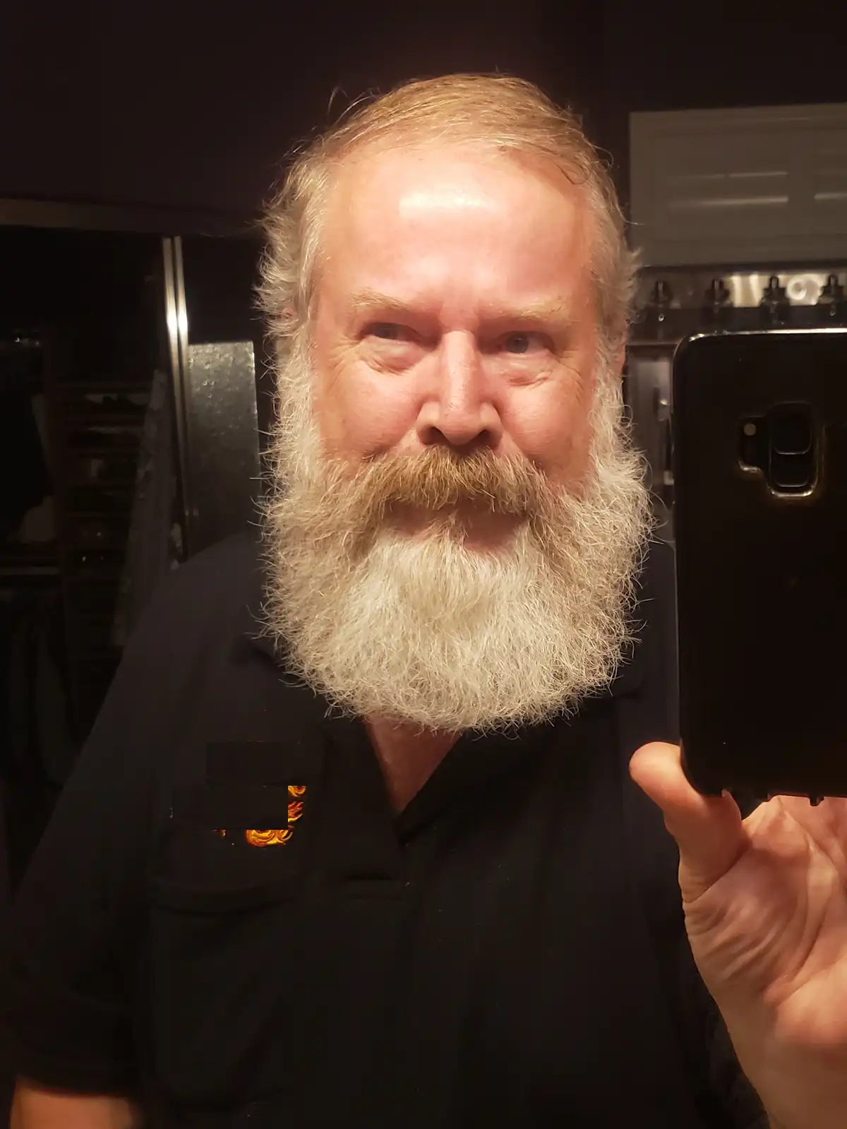 A middle-aged man with a long, full white beard and mustache takes a selfie in a mirror while wearing a black polo shirt. He…